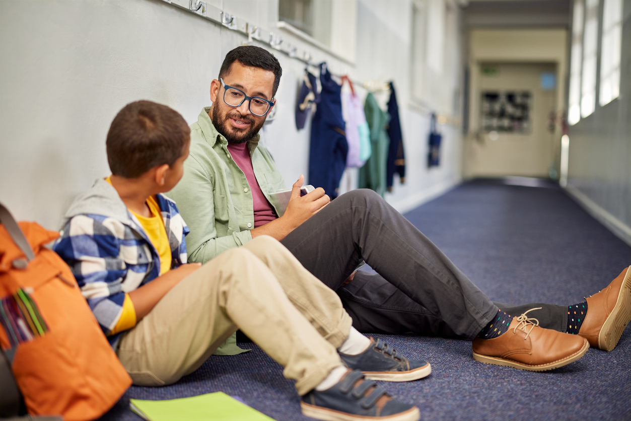 An adult sits beside a pupil and speaks with them in a school corridor.